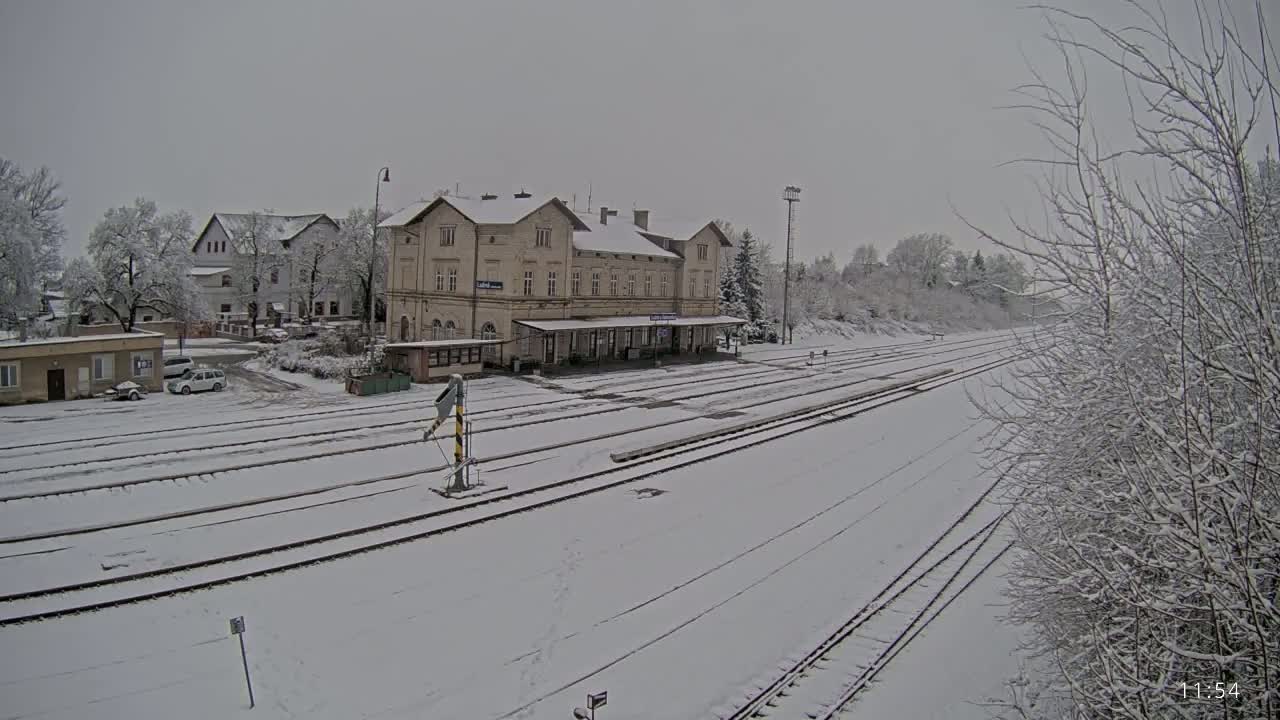 A snow-covered train station with several tracks and surrounding buildings is visible under an overcast sky.