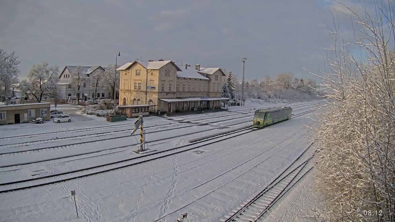 A light-green train is arriving at a snow-covered train station, with several buildings and trees also covered in snow.
