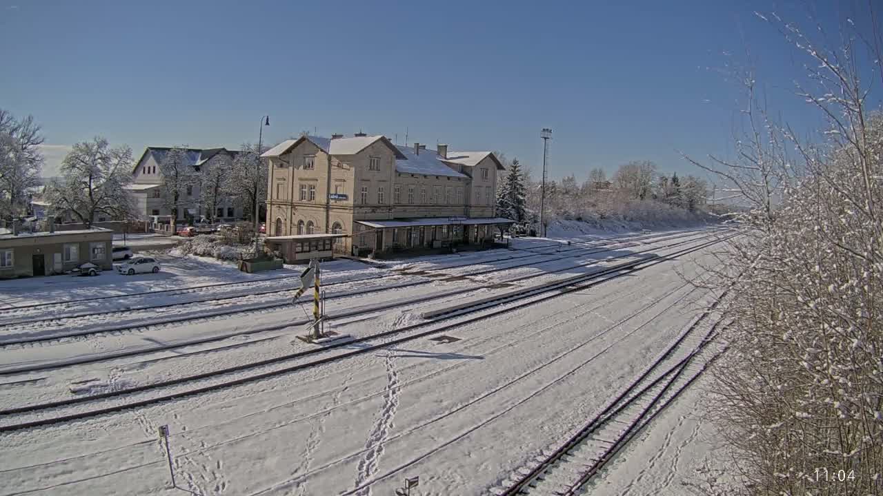 A snow-covered train station with several tracks and surrounding buildings under a clear, sunny sky.