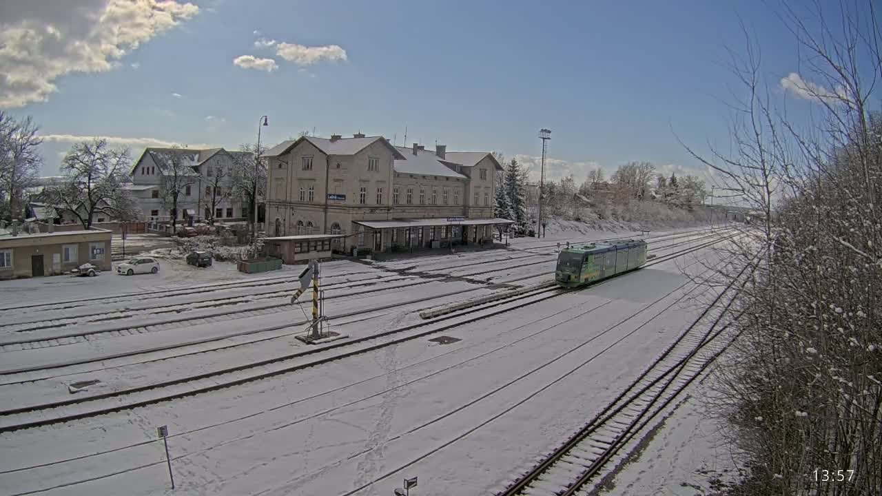 A light-green train approaches a snow-covered train station on a partly sunny day.