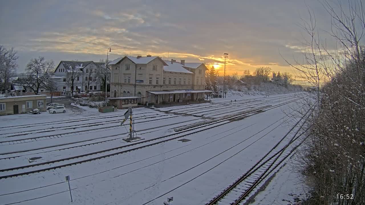 A snow-covered train station with several tracks and a long platform is bathed in the golden light of a setting sun.