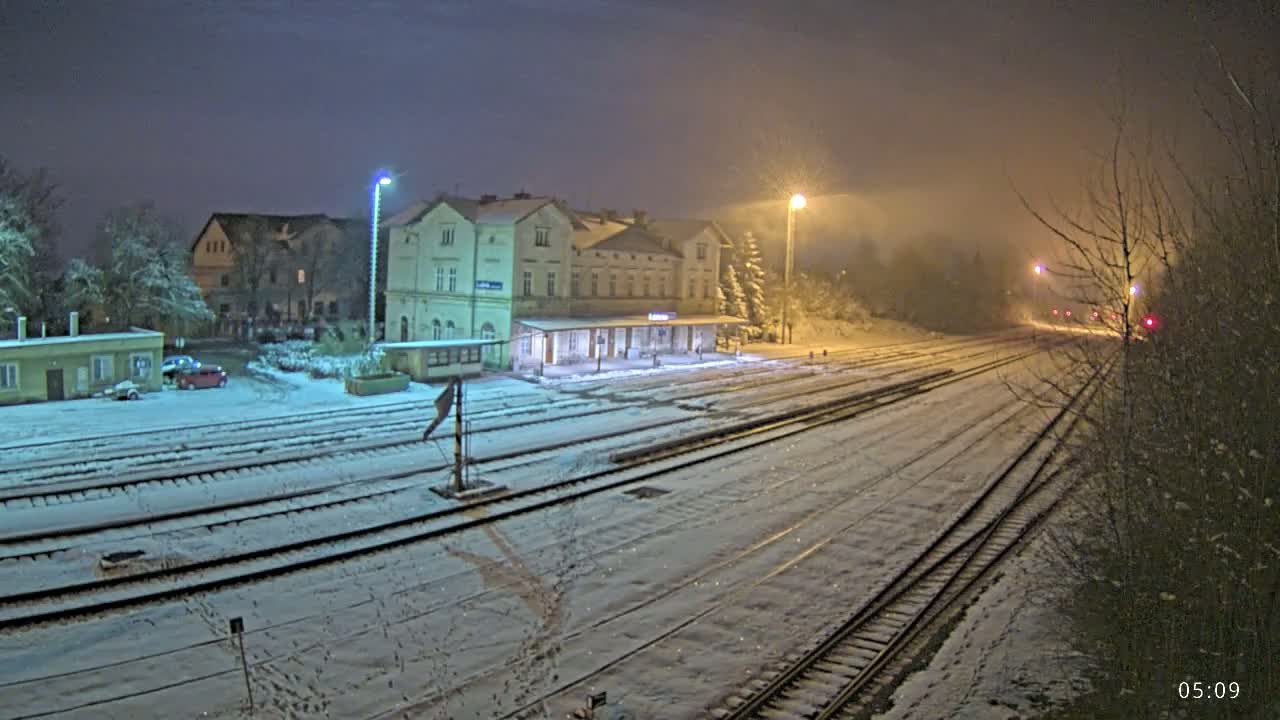 A snow-covered train station and tracks are illuminated at night under a hazy sky.