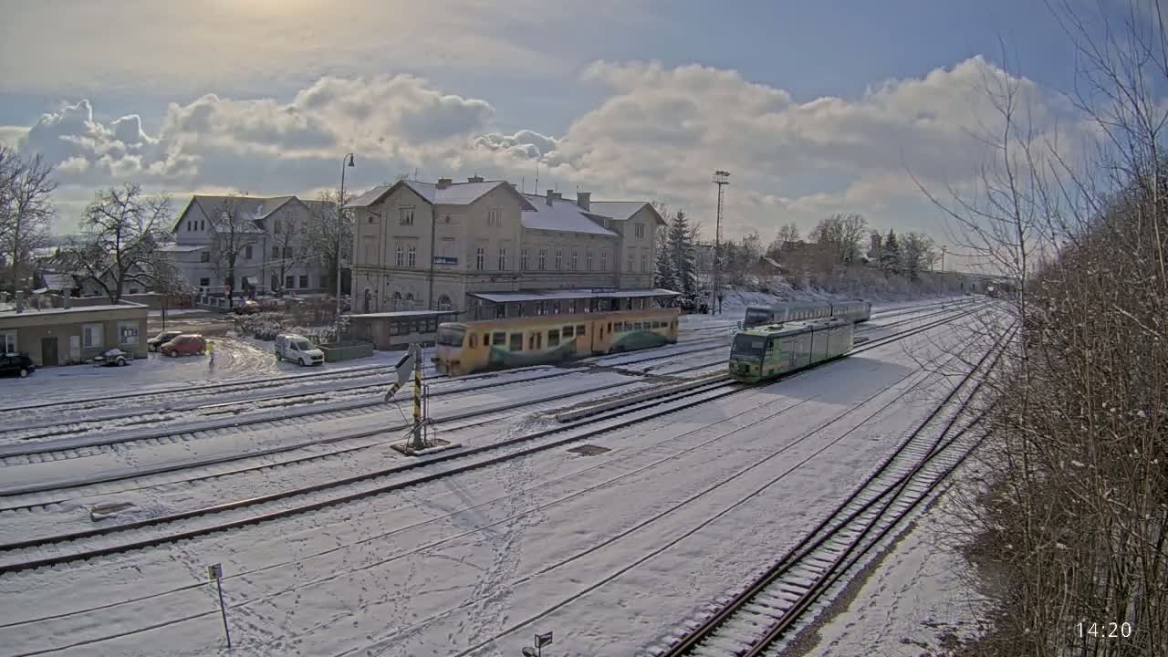 Two trains are at a snow-covered train station in front of a large building under a partly cloudy sky.