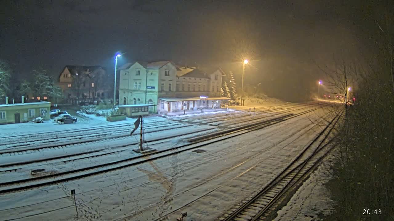 A snow-covered train station at night, illuminated by several lights, with several train tracks extending into the distance.