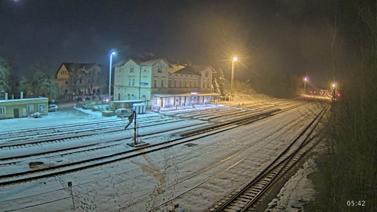 A snow-covered train station at night, illuminated by several streetlights, with train tracks extending into the distance.