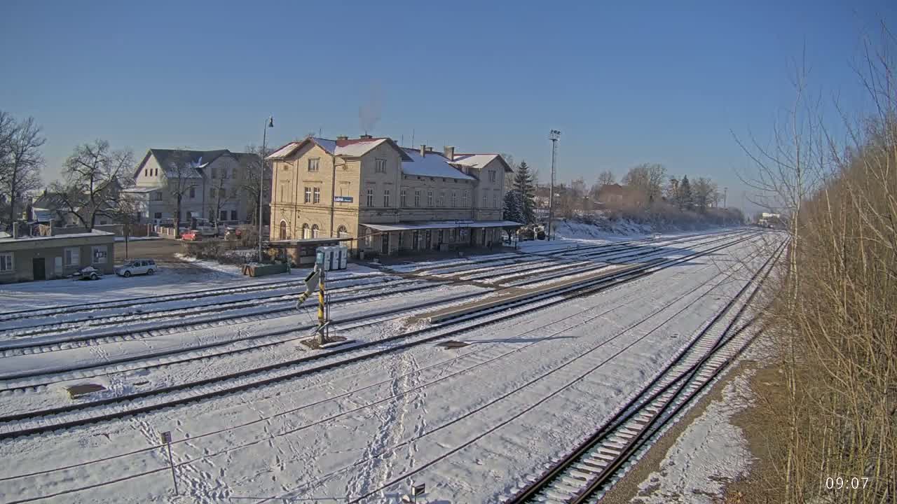A snow-covered train station with multiple tracks and several buildings in the background under a clear blue sky.