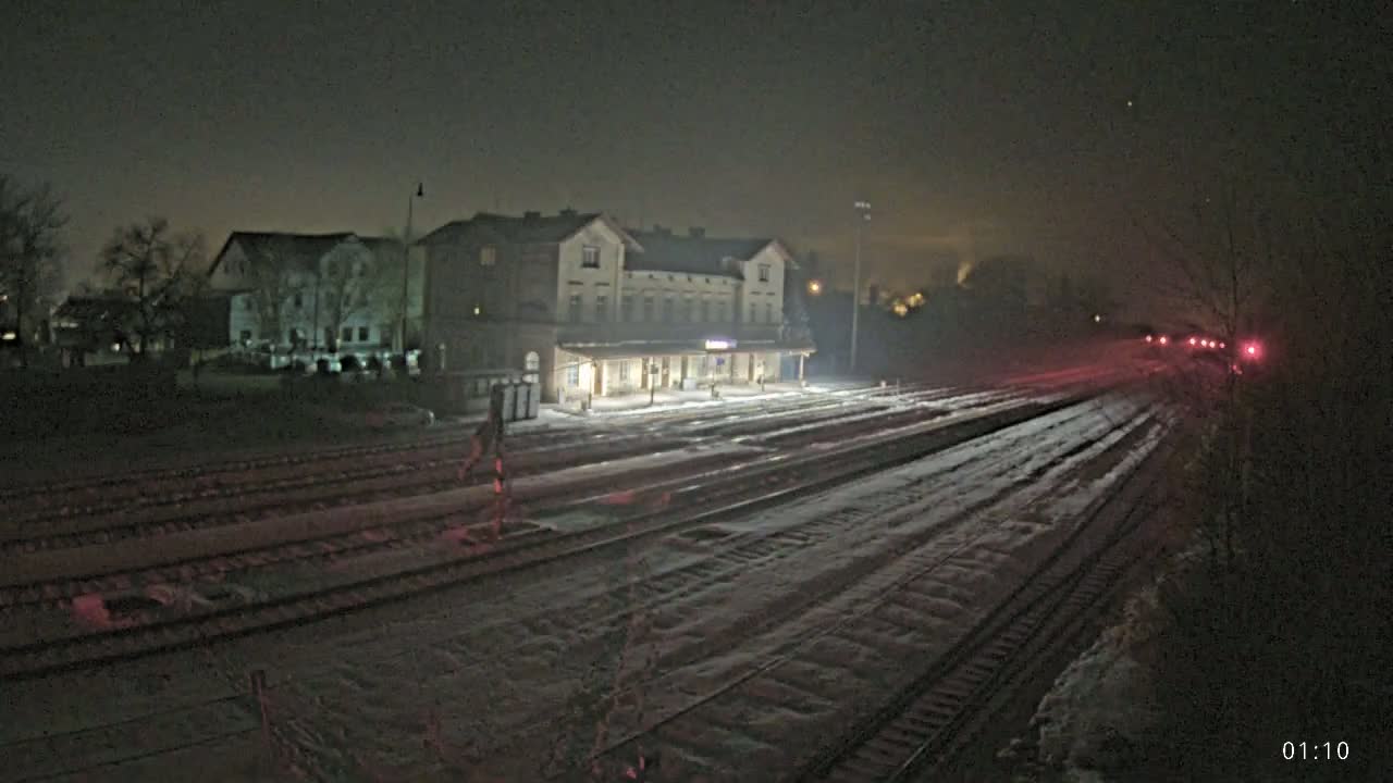 A snow-covered train station and tracks are illuminated at night under a dark, hazy sky.