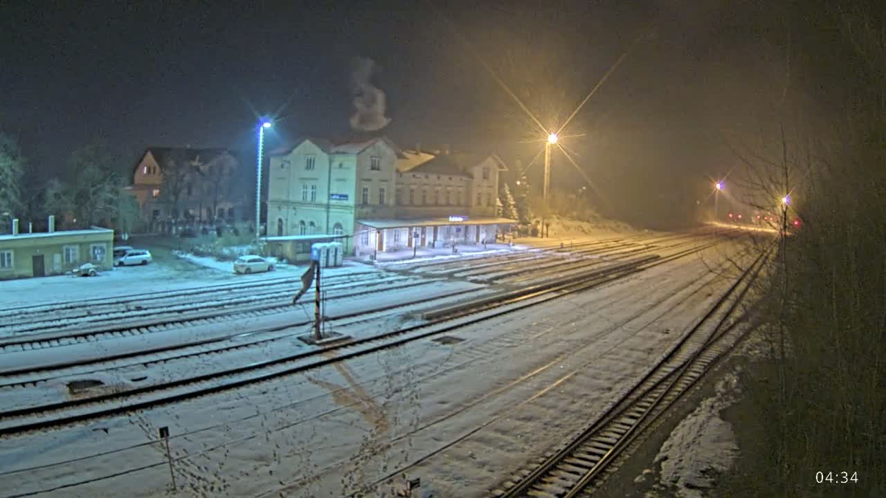 A snow-covered train station at night, illuminated by streetlights, with several train tracks extending into the distance.