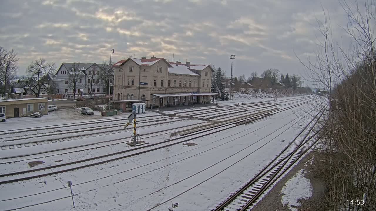 A snow-covered train station with multiple tracks and several buildings in the background under a cloudy sky.