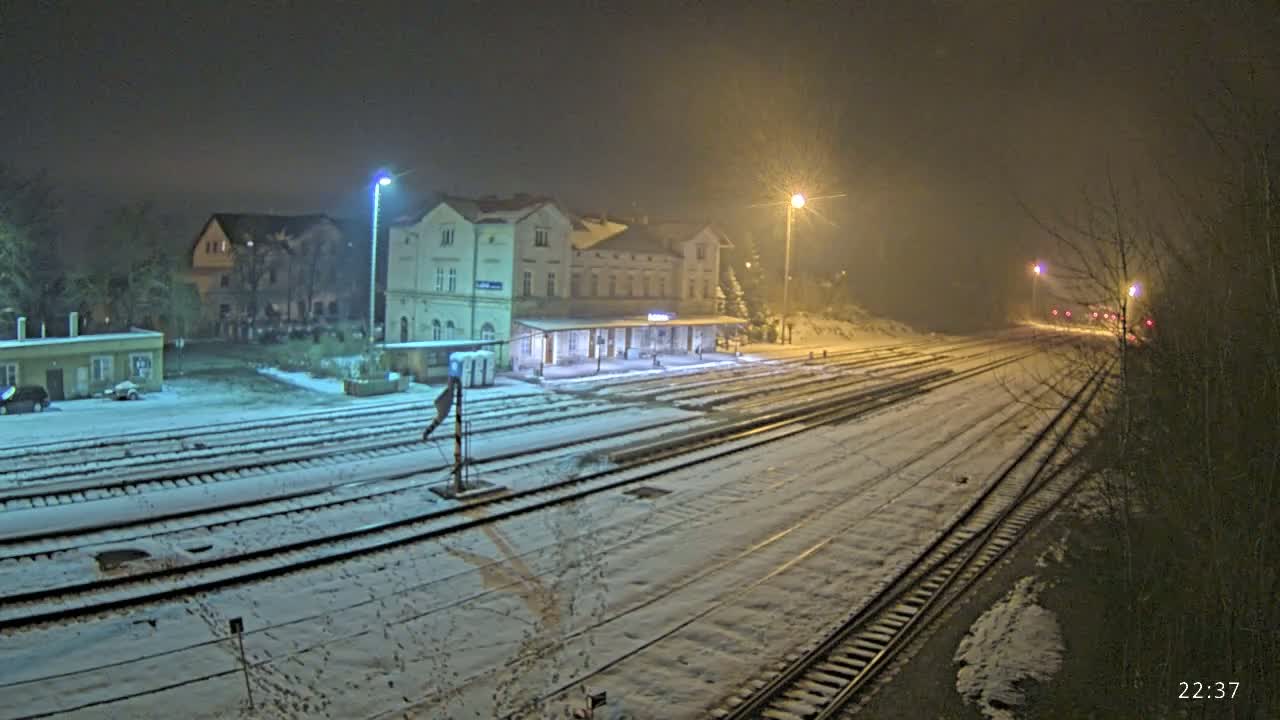 A snow-covered train station and tracks are illuminated at night by streetlights under hazy conditions.