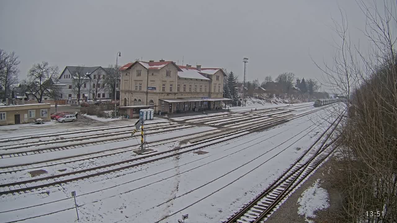 A snow-covered train station with multiple tracks and a long building, situated in a town under an overcast sky.