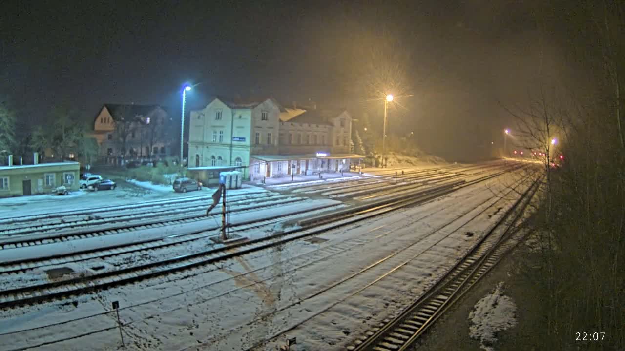 A snow-covered train station at night, illuminated by streetlights, with several cars parked nearby.