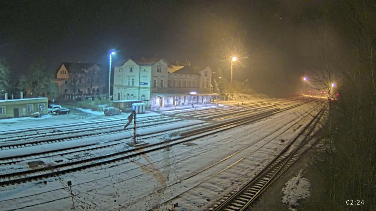 A snow-covered train station at night, illuminated by several lights, with multiple train tracks extending into the distance.