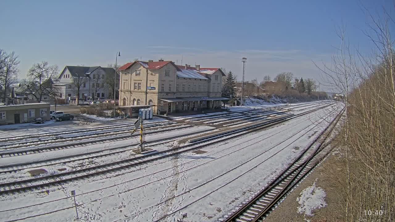 A snow-covered train station with multiple tracks extends into the distance under a sunny sky.