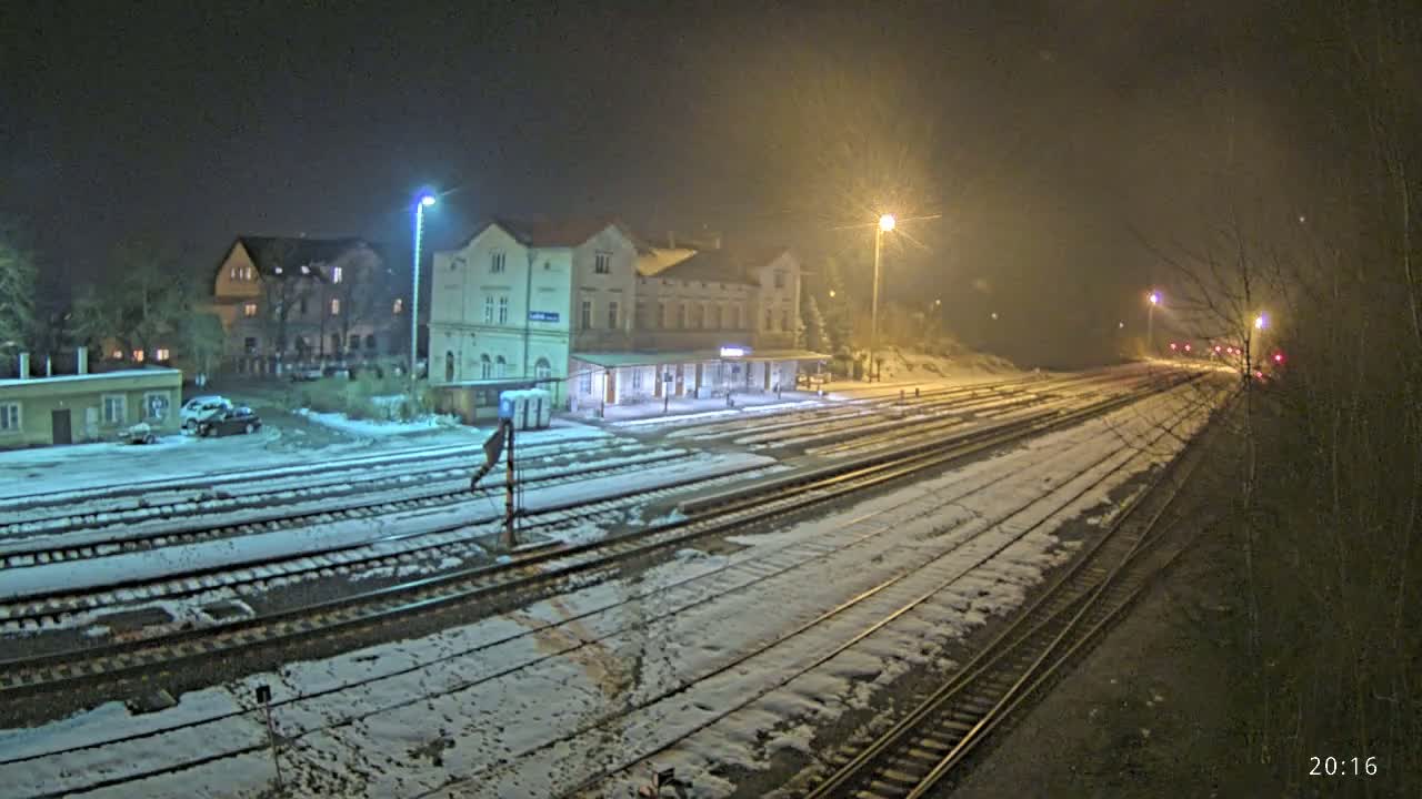 A snow-covered train station at night, illuminated by streetlights, with multiple train tracks extending into the distance.