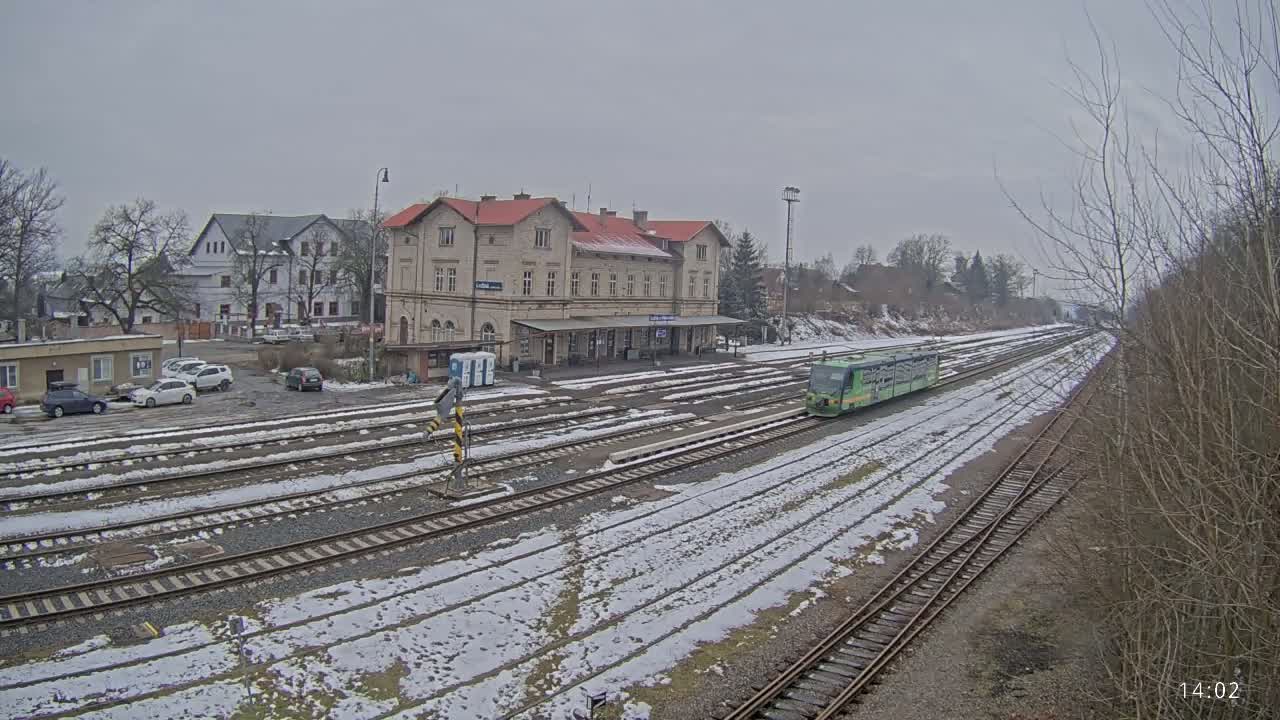 A light-green train is arriving at a snow-covered train station with a long building and a parking lot, under an overcast sky.