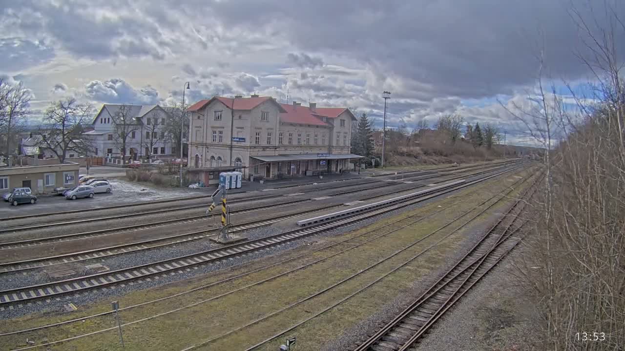 A train station with a long platform and several tracks is visible under a cloudy sky, with a parking lot and other buildings in the background.