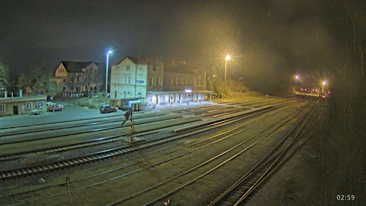 A nighttime view of a train station with several tracks and a few cars parked in the lot, under hazy light conditions.