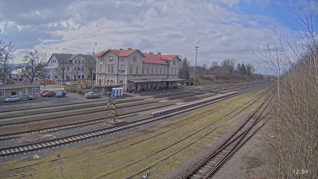 A long view of a train station with a long, light-colored building and several railway tracks under a partly cloudy sky.
