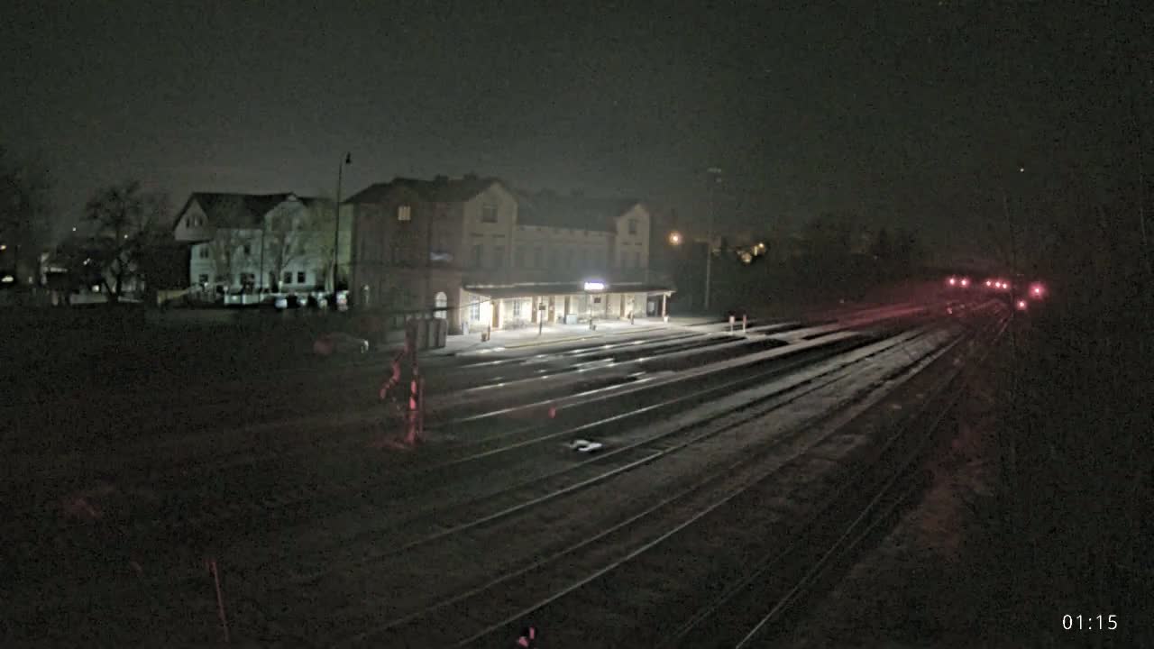 A brightly lit train station at night is seen from across a set of train tracks, with a few houses visible in the background under a dark sky.