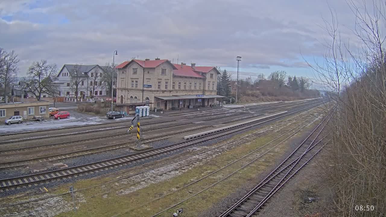 A train station with a long platform and several tracks is visible under an overcast sky.