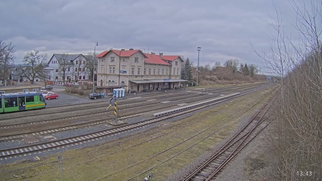 A long view of a train station with a light-green tram and several cars in the parking lot under an overcast sky.