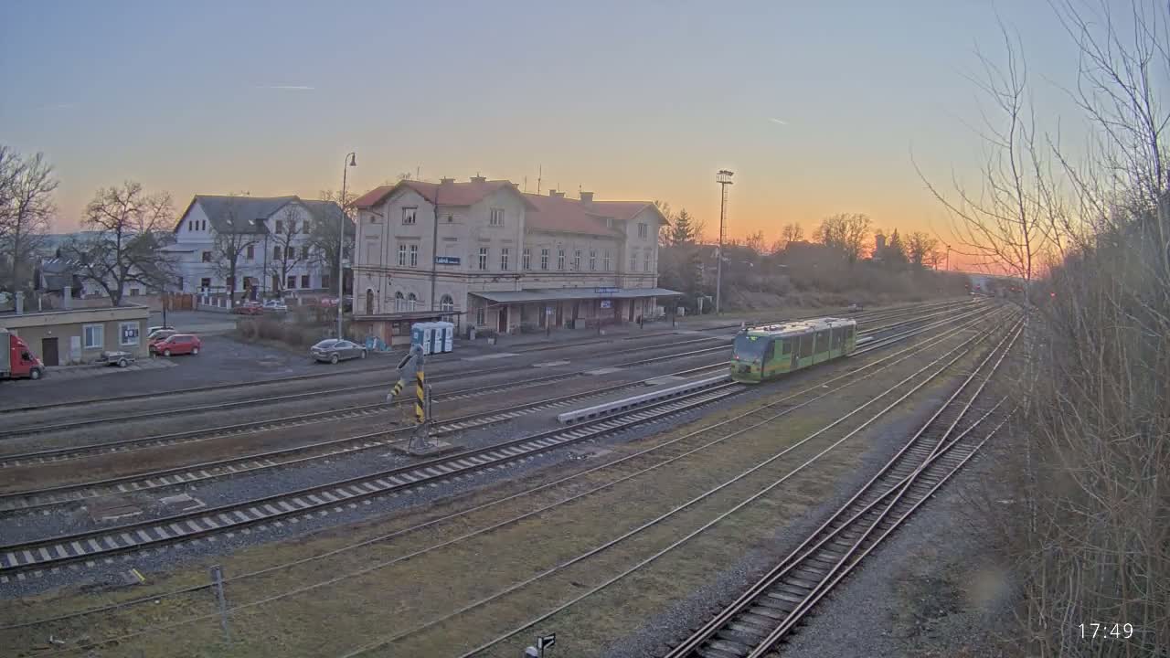 A light-green train is at a train station during sunset, with several train tracks and a few cars parked in a lot nearby.