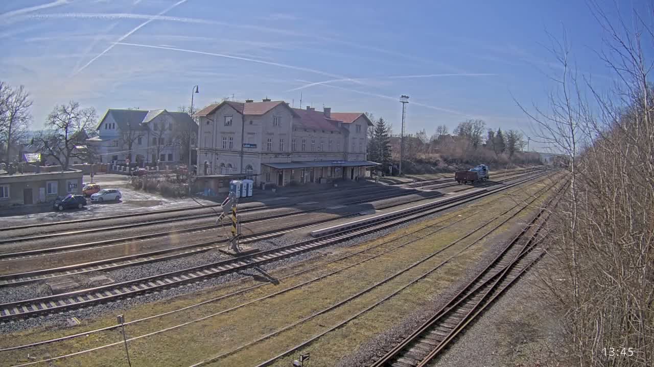 A train station with a long building and multiple train tracks is shown on a sunny day.