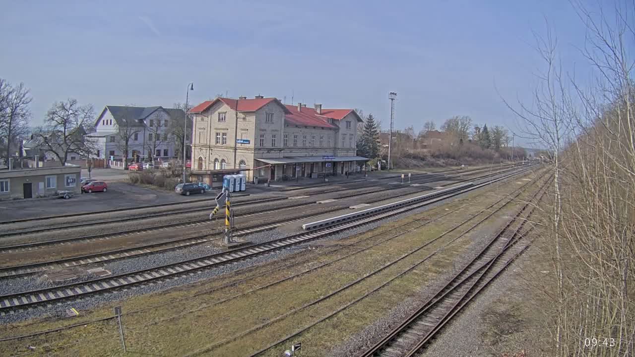 A long view of a train station with a large building and multiple train tracks under a clear, sunny sky.
