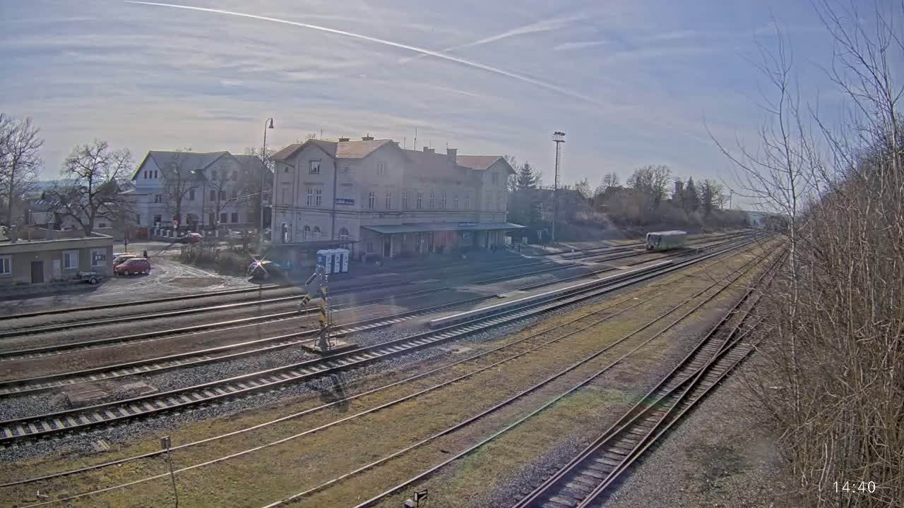 A train station with a long platform and several tracks is visible under a sunny sky, with a single train car in the distance.