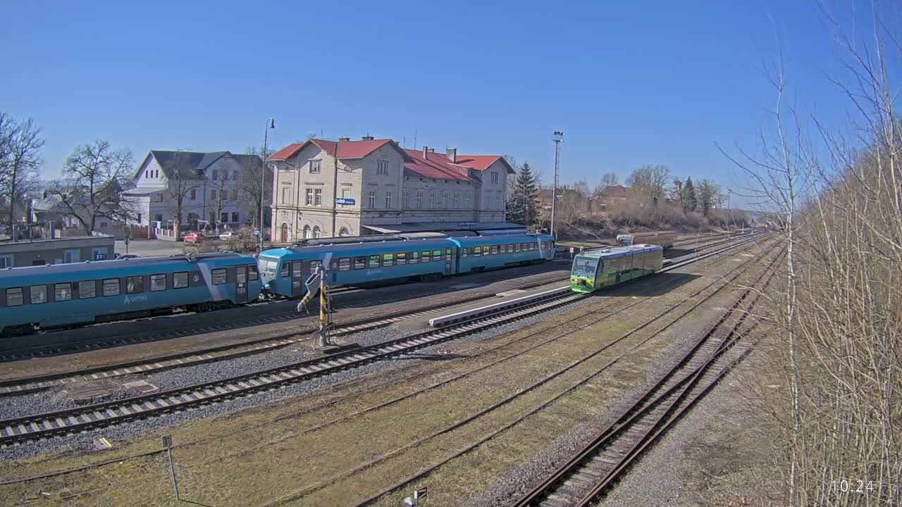 Under a clear blue sky, two teal trains and a lime green train are parked at a train station in front of a large building.