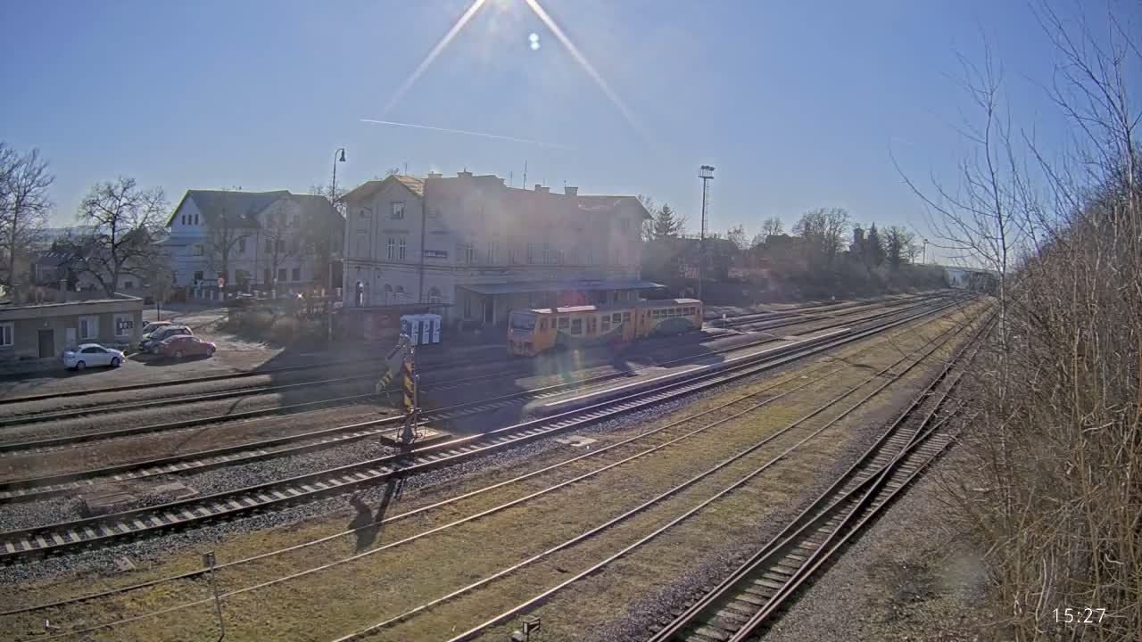 A train sits at a station on a sunny day next to several train tracks and a parking lot with cars.