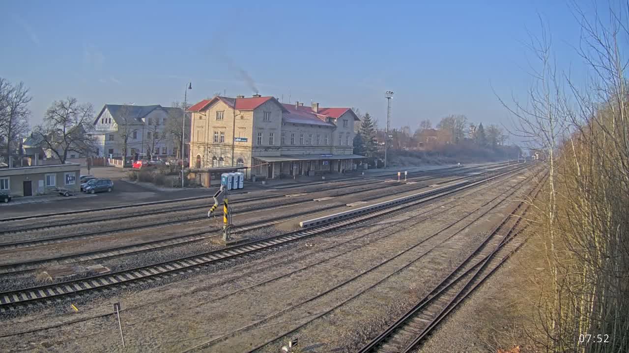 A long view of a train station on a clear, sunny day shows several train tracks extending to the horizon, with a large light-colored building in the center and other buildings in the background.
