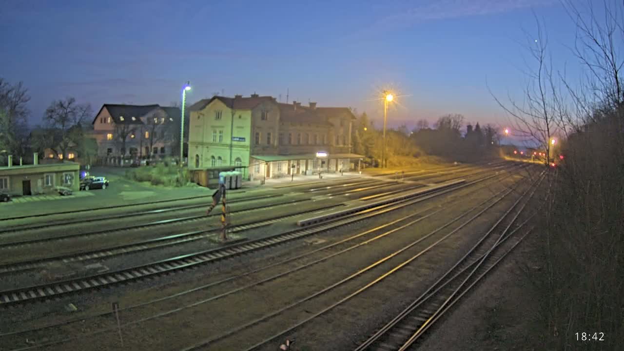 A train station with a long platform and several sets of tracks is illuminated at dusk under a twilight sky.