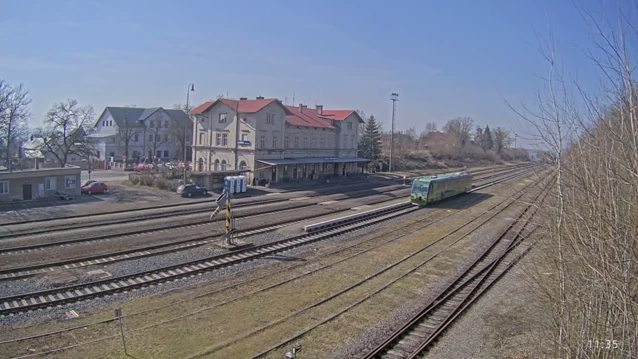 A light-green train sits at a train station on a sunny day, with several tracks leading away from a long, light-colored building.