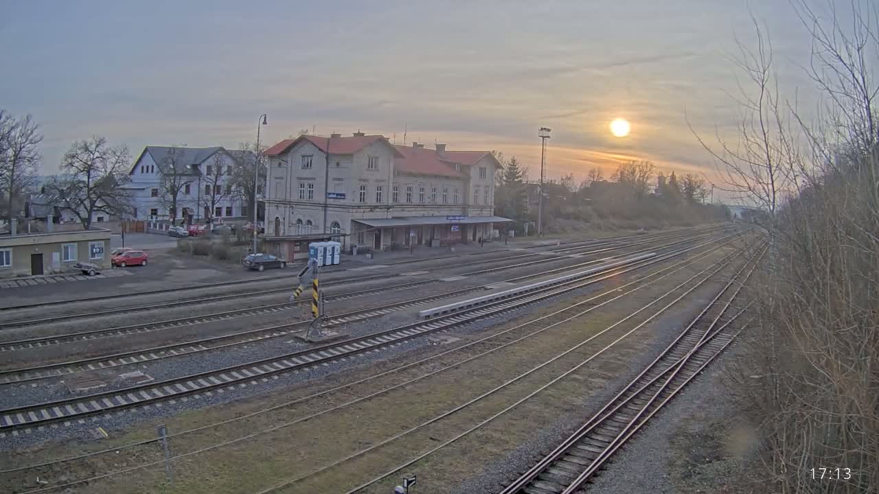 A train station with a long platform and multiple tracks is visible at sunset, with a few cars parked nearby.