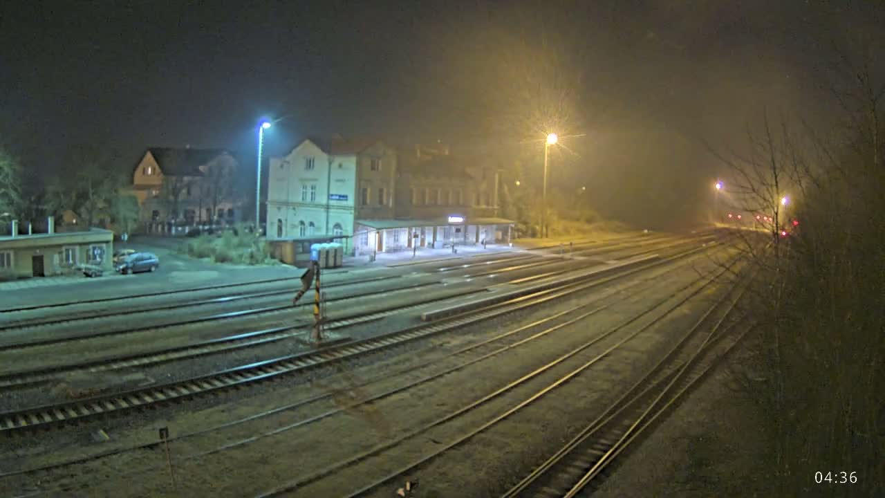 A nighttime view of a train station with several tracks extending into the foggy distance, illuminated by streetlights.