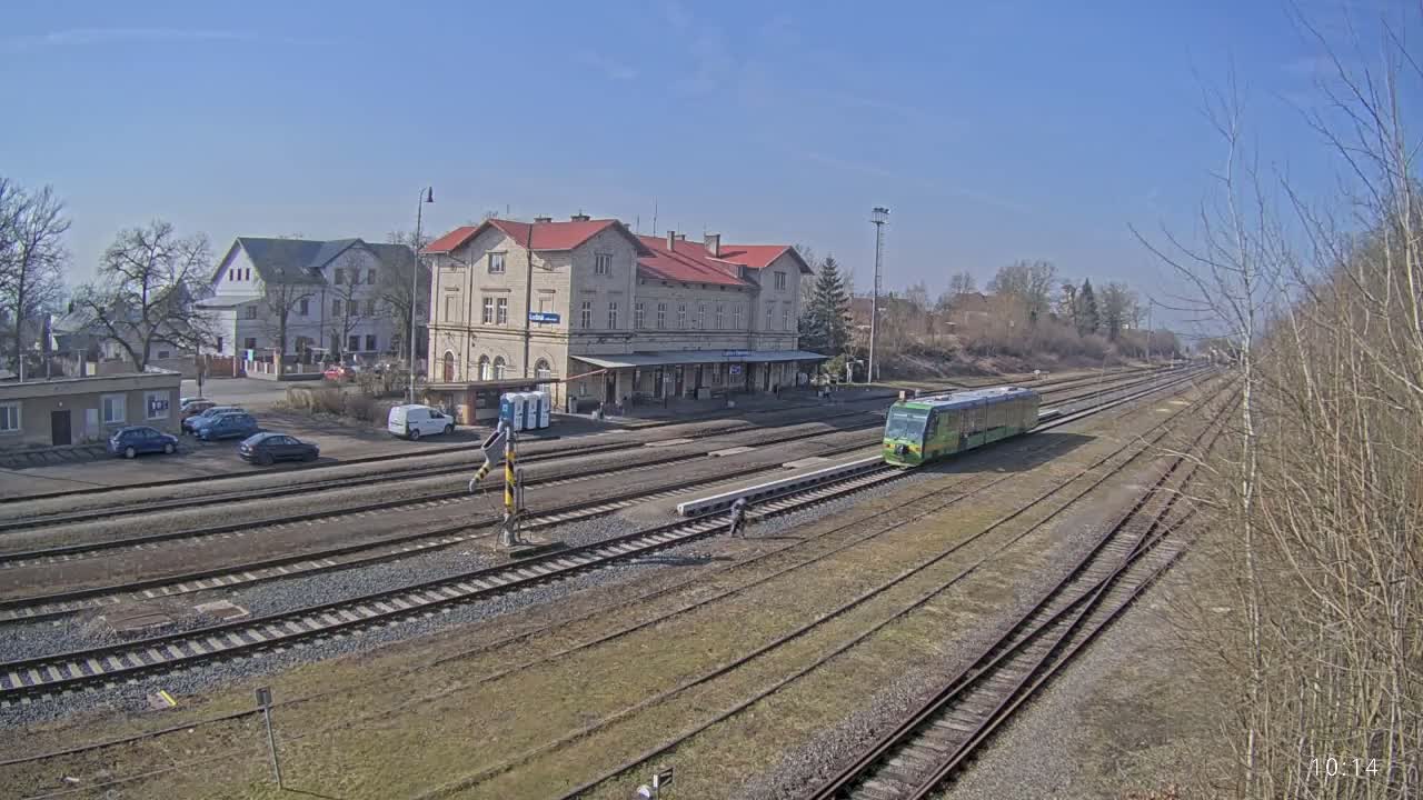 A light-green train is arriving at a train station on a sunny day, with a large tan building serving as the station house.