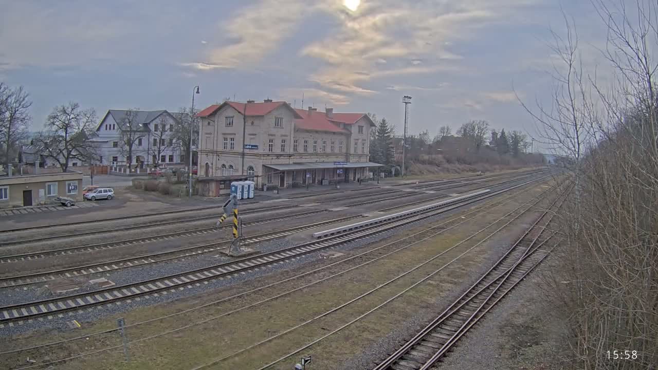 A train station with a long platform and multiple tracks is situated under an overcast sky, surrounded by buildings and bare winter trees.