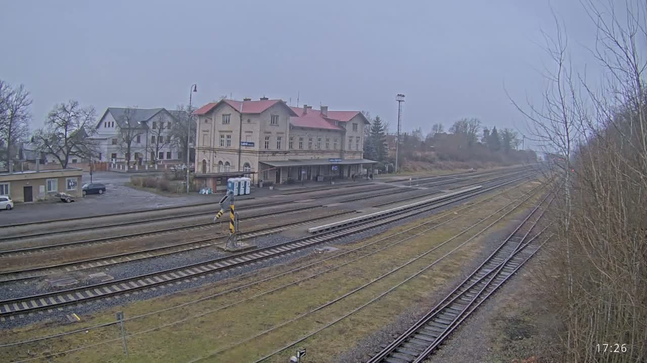 A long view of a train station on an overcast day shows multiple train tracks extending into the distance, with a two-story station building in the foreground and other buildings visible in the background.
