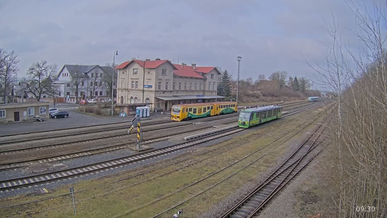 Two trains are stopped at a train station on an overcast day, next to a long, low building.