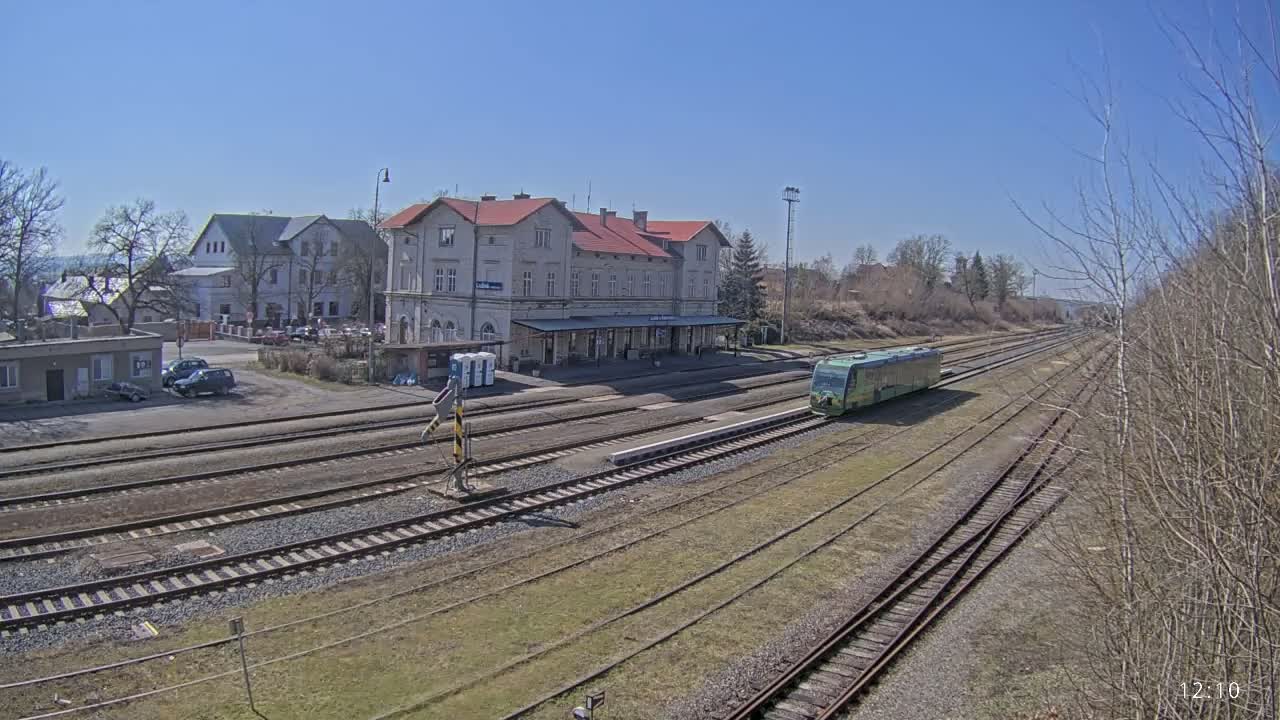 A light-green train sits on a railway track next to a long building with a red roof, in front of a small town under a clear blue sky.