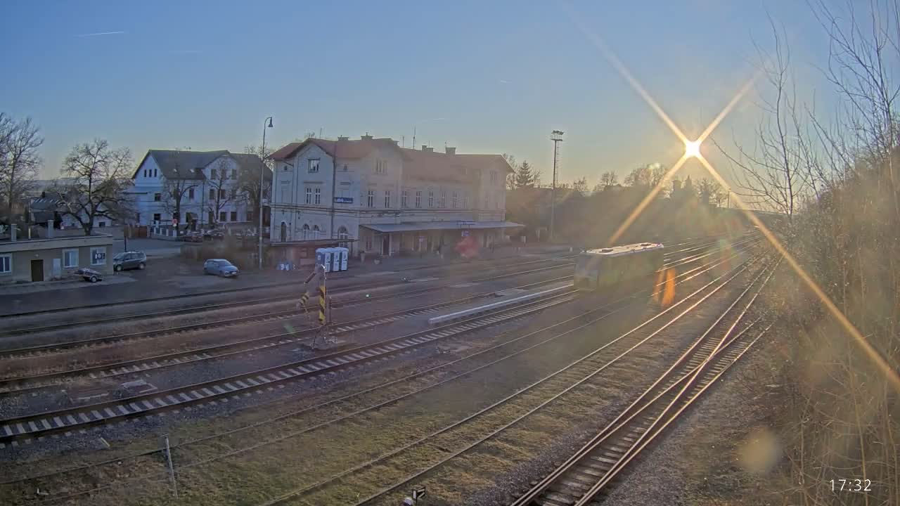 A train sits on the tracks at a train station in the late afternoon sun, with several buildings visible in the background.