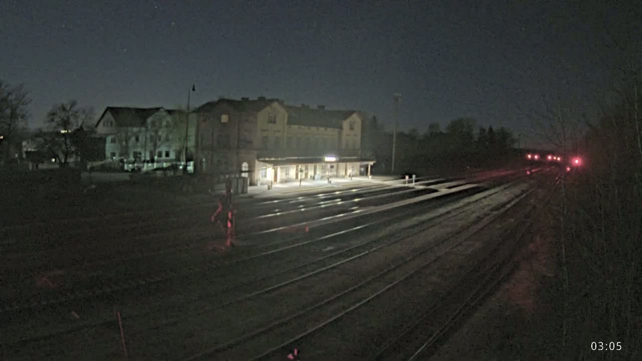 Under a dark, clear night sky, a train station with illuminated platforms sits beside several train tracks leading into the distance, where distant red lights are visible.