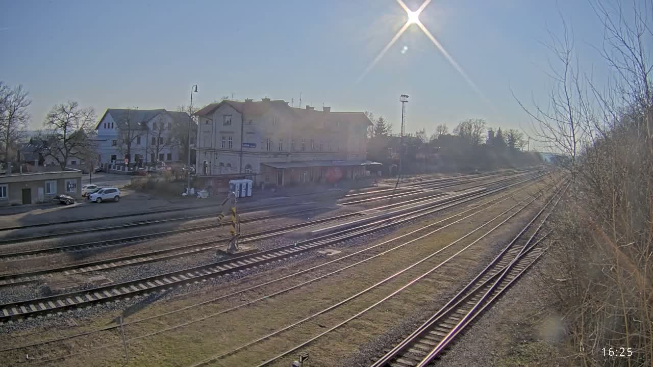A sunny day reveals a train station with multiple tracks extending into the distance, flanked by buildings and bare trees.