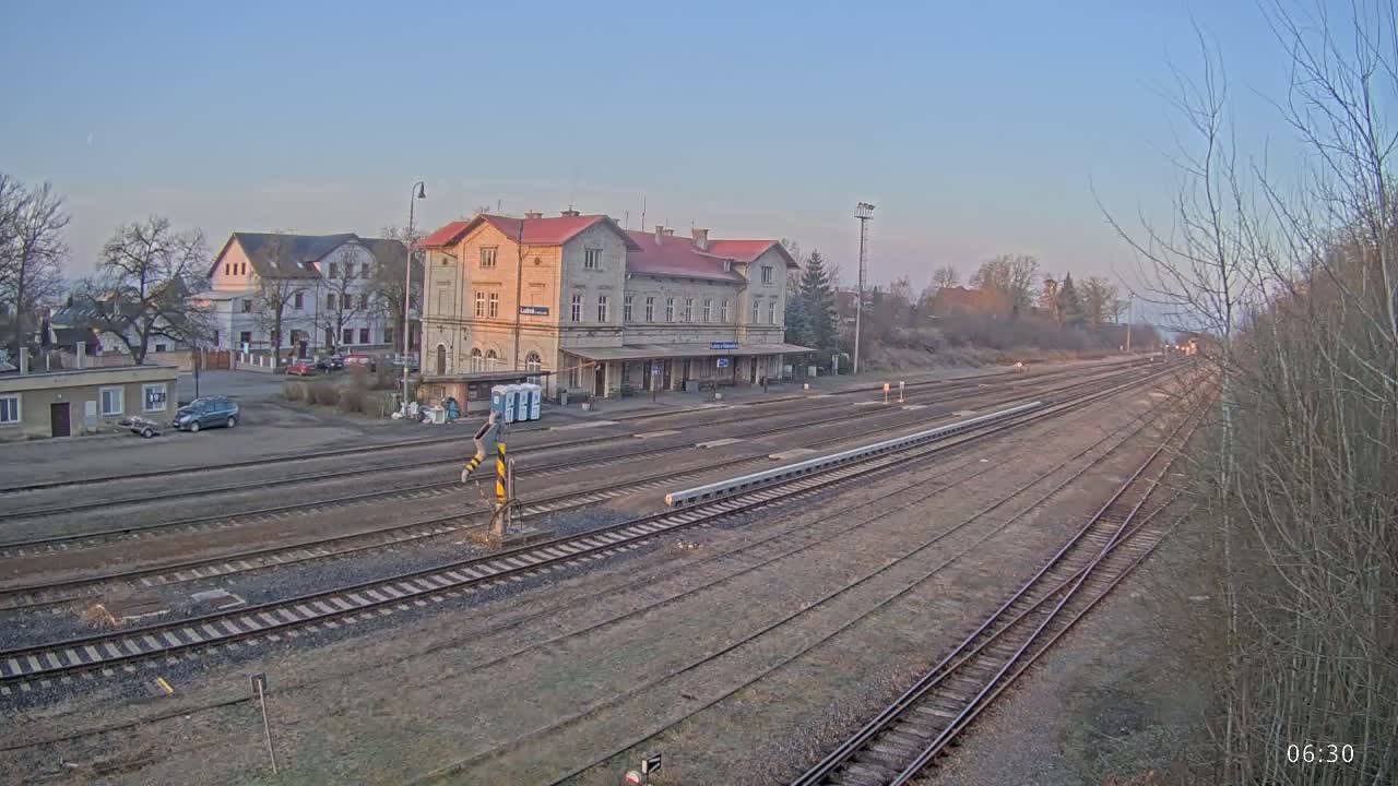 A long view of a train station with a large, light-colored building, multiple train tracks extending into the distance, and bare trees under a clear, early morning sky.