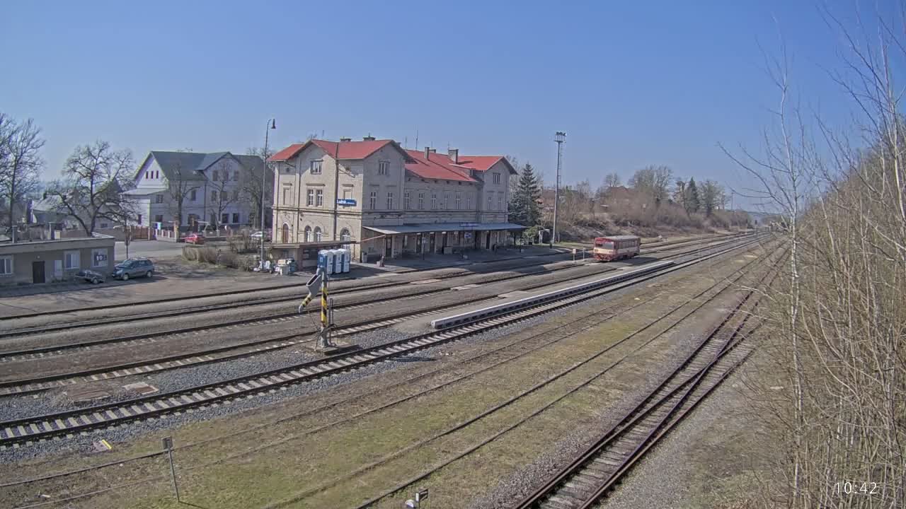 A train station with a long platform and several train tracks is visible on a sunny day.