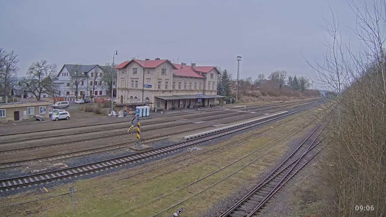 A train station with a long platform and multiple train tracks is visible, under an overcast sky.