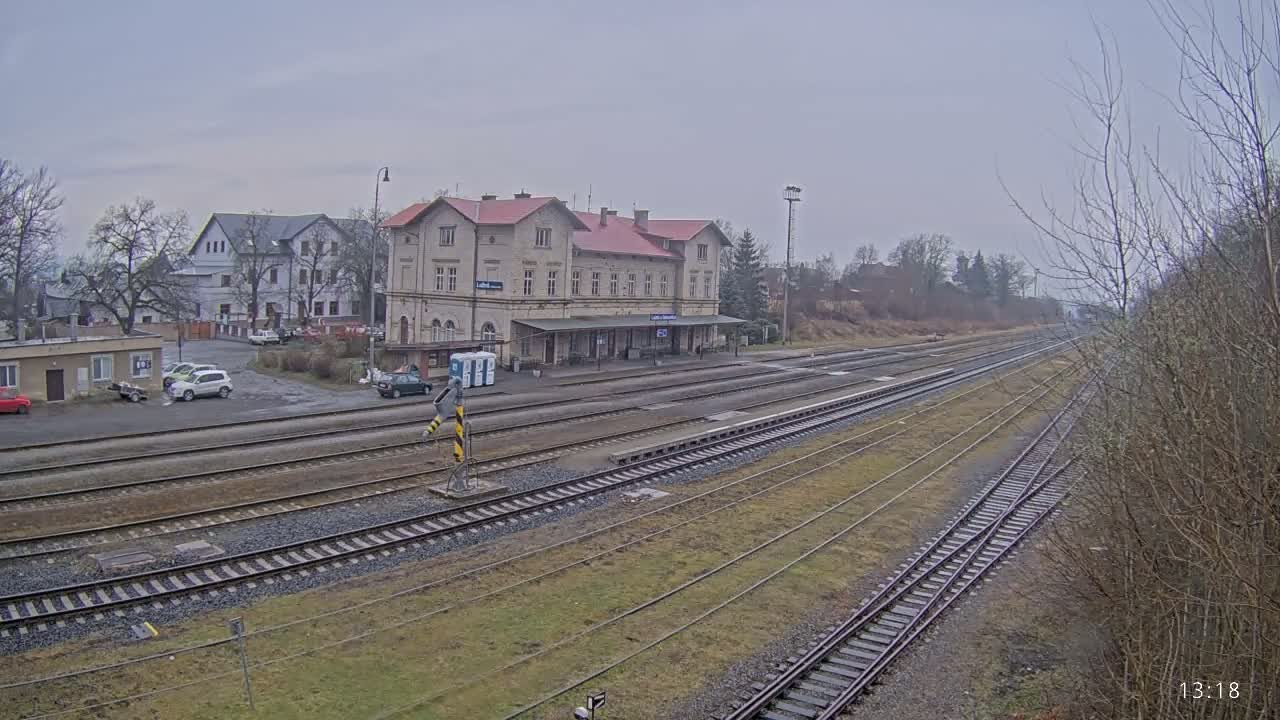A train station with a long building and several tracks stretches into the overcast distance, with a small parking lot and other buildings visible nearby.