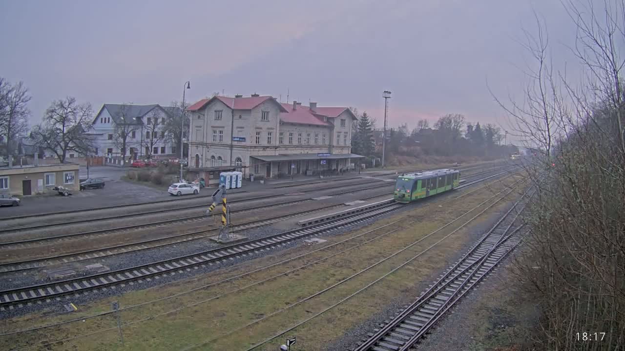 A light-green train is leaving a train station on an overcast day, passing by a large, light-colored building.